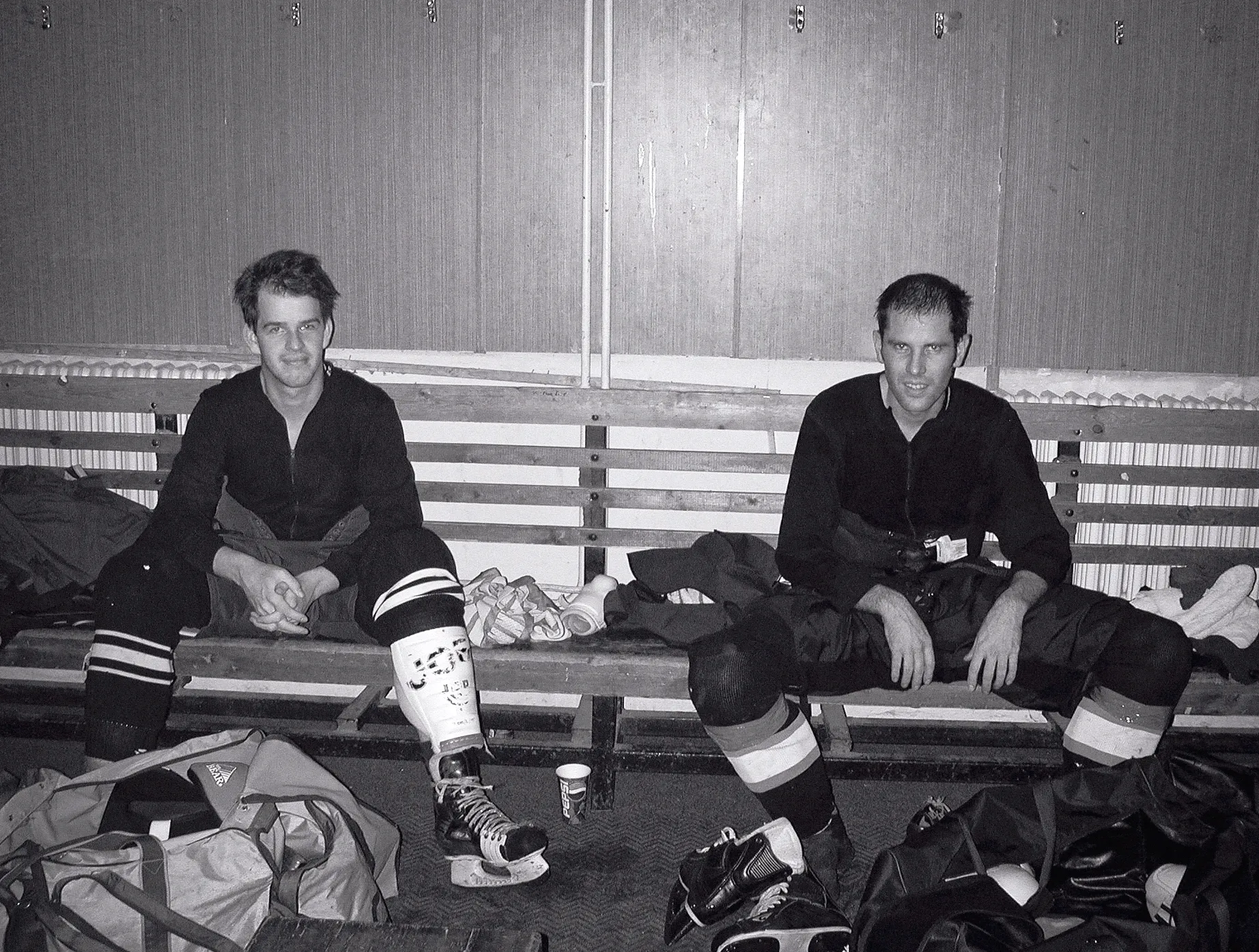 Dos jugadores de hockey sobre hielo con equipo completo sentados en un banco de madera en un vestuario, rodeados de bolsas deportivas y patines.