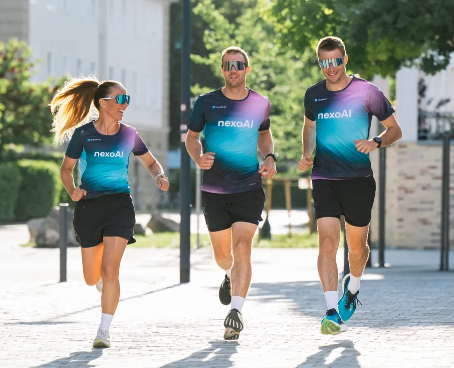 Tres atletas, dos hombres y una mujer, corren con camisetas de carrera coloridas a juego y pantalones cortos en un camino pavimentado.