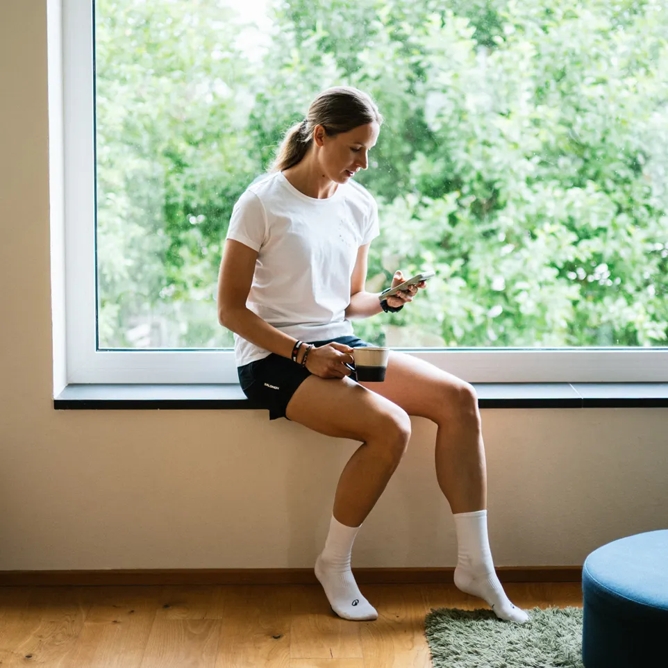 Una mujer está sentada en un alféizar de ventana, vistiendo una camiseta blanca y pantalones cortos negros, mientras sostiene un teléfono inteligente y disfruta de una taza de café.