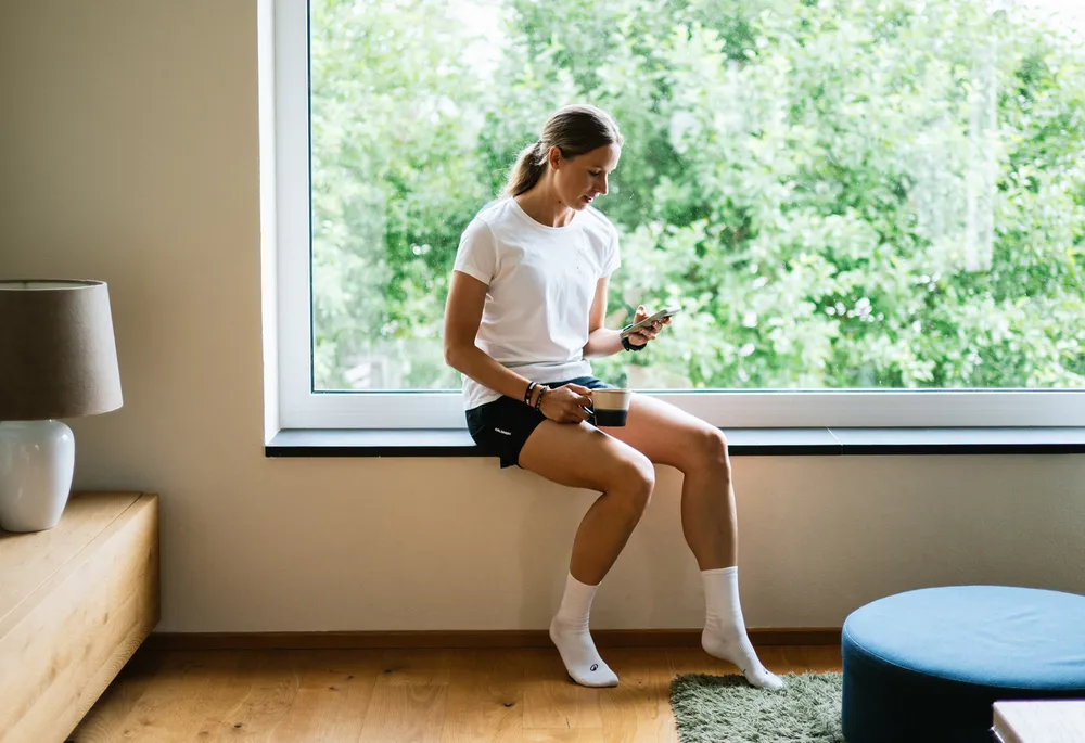 Una mujer está sentada en el alféizar de una ventana sosteniendo un teléfono en una mano y una taza en la otra, vestida con una camiseta blanca y pantalones cortos negros.
