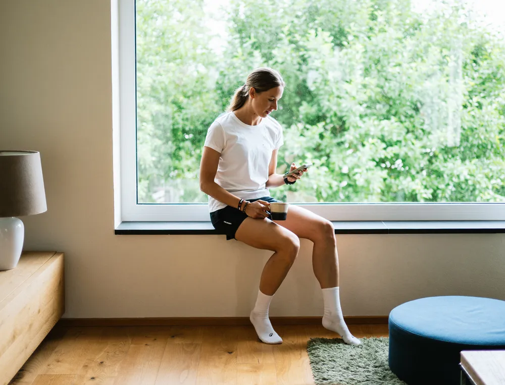 Una mujer está sentada en un alféizar de ventana, mirando su teléfono mientras sostiene una taza de café, vestida con una camiseta blanca y pantalones cortos negros.