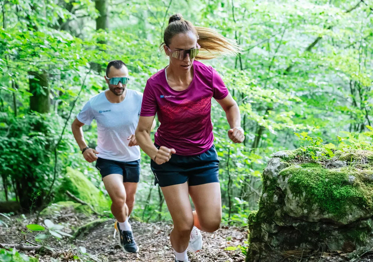 Dos corredores, un hombre con una camiseta blanca y una mujer con una camiseta deportiva roja, corren por un sendero boscoso.