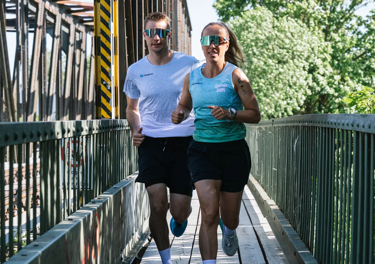 Un hombre y una mujer están corriendo en un puente, ambos visten ropa deportiva y gafas de sol.