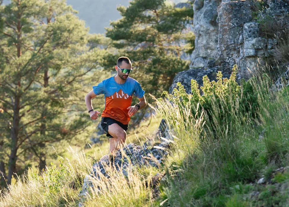 Un corredor con una camiseta colorida y pantalones cortos corriendo por un camino rocoso en un paisaje boscoso verde.