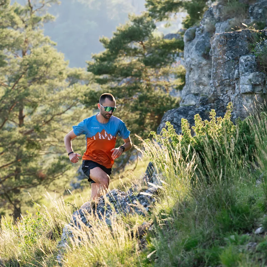 Un corredor con una camiseta deportiva azul y naranja corriendo por un camino rocoso en un paisaje boscoso soleado.