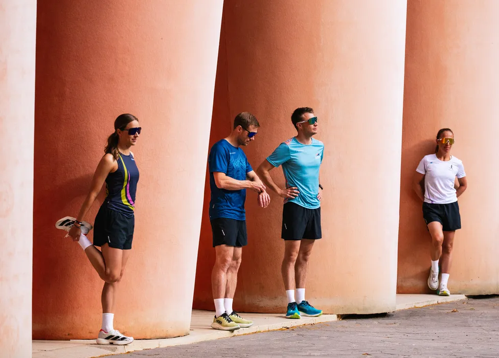 Un grupo de cuatro atletas en ropa de correr se estira contra una pared con grandes columnas.