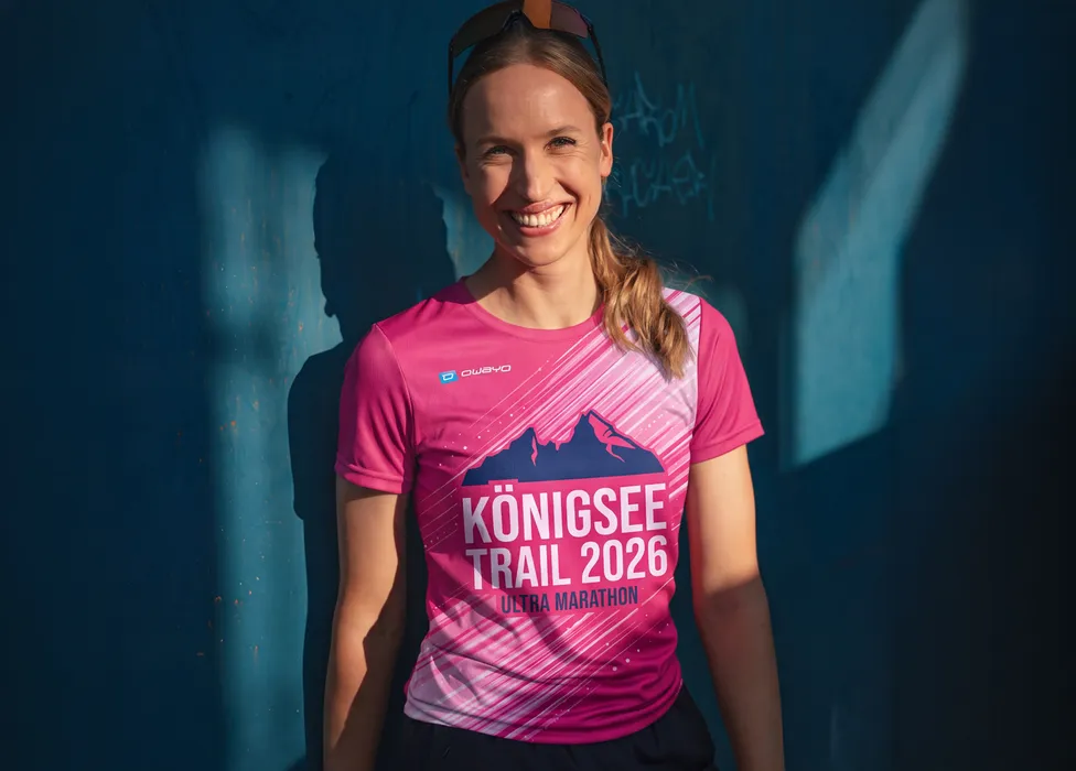 Una mujer sonriente lleva una camiseta rosa con la impresión 'Königsee Trail 2026 Ultra Marathon' frente a una pared azul.
