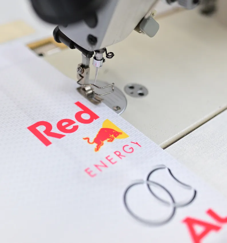Una máquina de coser está cosiendo una prenda deportiva blanca con el logotipo rojo de Red Bull.
