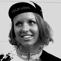 Una mujer con cabello rubio corto lleva una gorra de ciclismo negra con la inscripción 'VELO LOVE' y un maillot de ciclismo personalizado con el nombre 'Anja Sturm'.