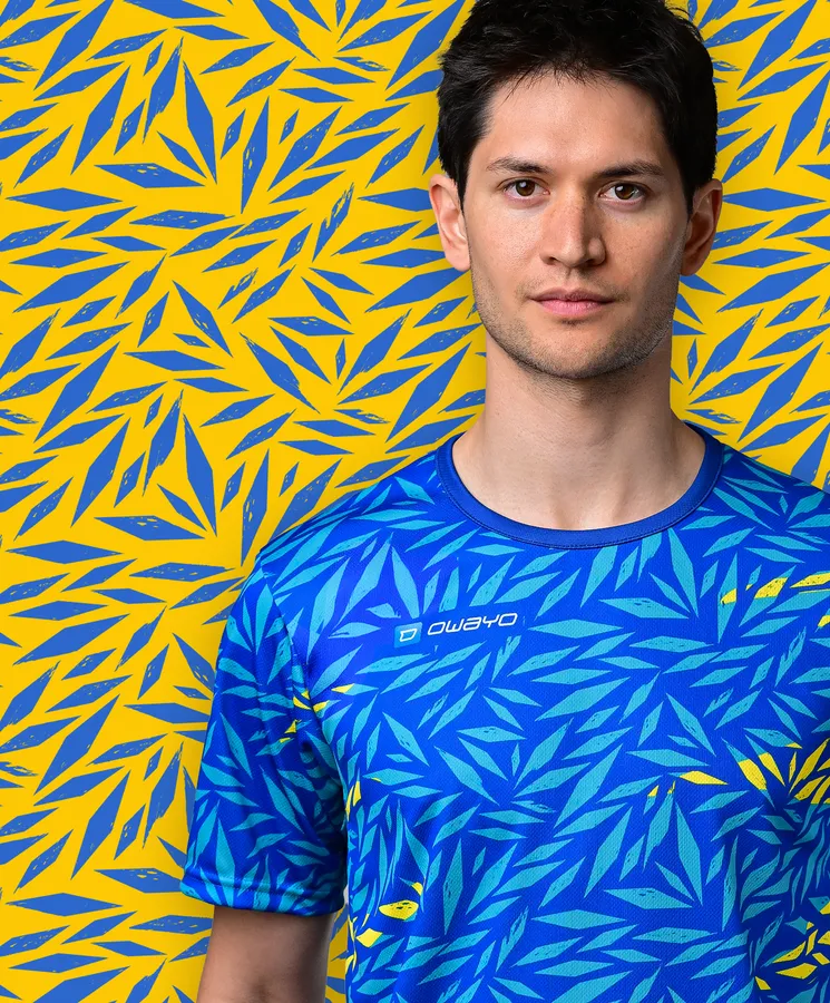 Un joven lleva una camiseta deportiva azul con un patrón de hojas coloridas sobre un fondo amarillo.