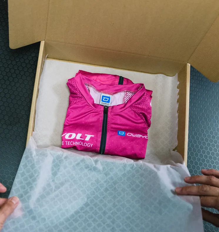 Una prenda deportiva personalizada de color rosa de owayo, doblada, está dentro de una caja de cartón abierta sobre papel blanco, sostenida por manos.