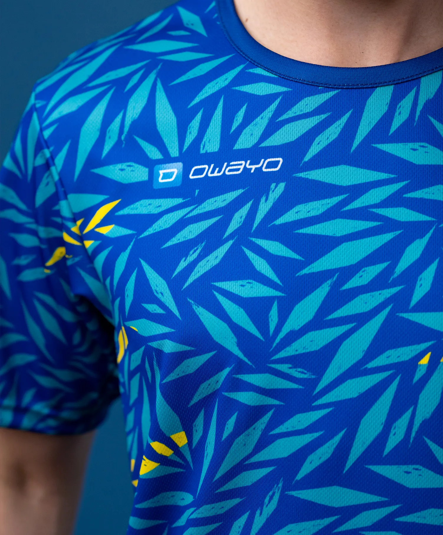 Una camiseta deportiva de manga corta con un patrón azul en forma de hojas en varios tonos de verde con acentos amarillos.