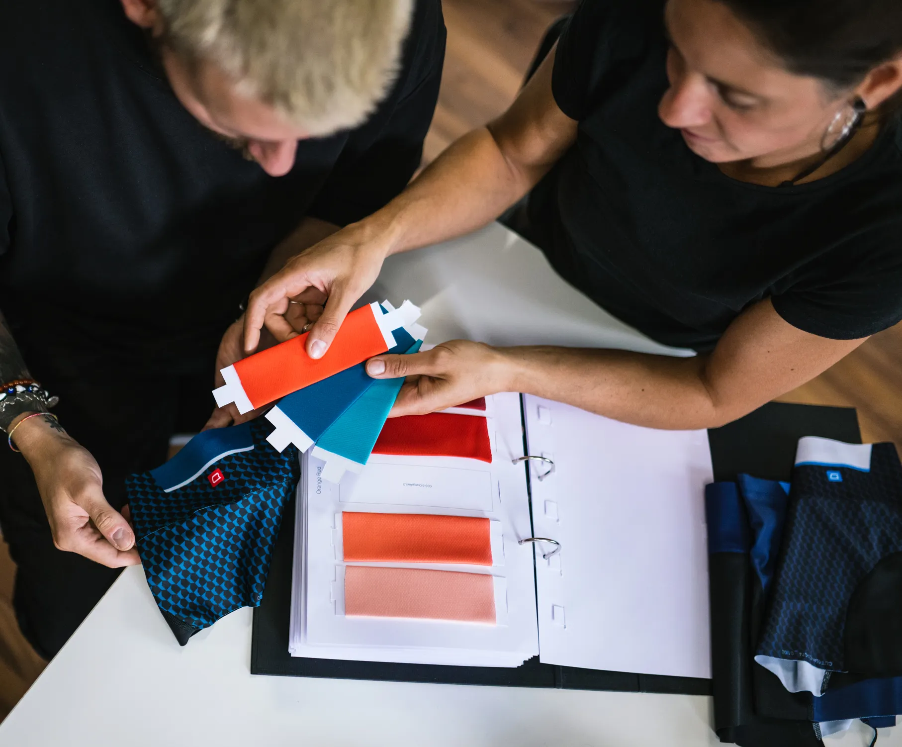 Dos personas seleccionan colores para ropa deportiva personalizada al examinar muestras de tela y un catálogo de colores.