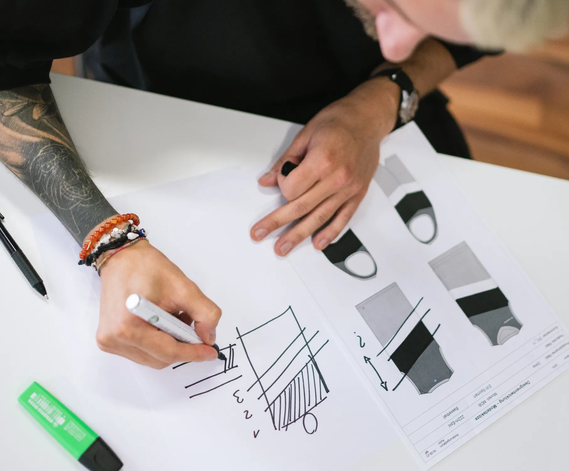 Una persona con un tatuaje en el brazo dibuja un diseño con un marcador en un papel, mientras que dibujos técnicos de ropa deportiva están sobre la mesa.