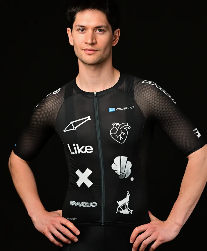 Un joven lleva una camiseta deportiva negra ajustada adornada con varios logotipos y diseños en blanco, posando frente a un fondo negro.