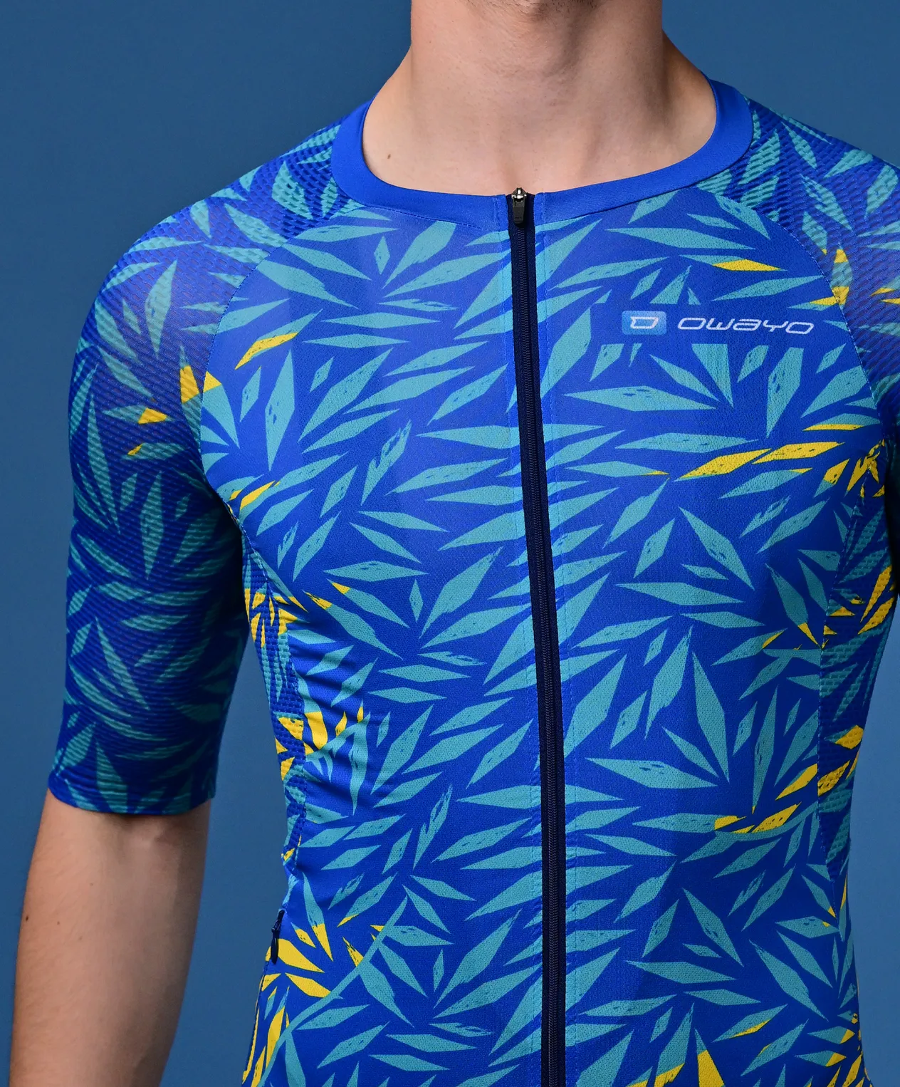 Un hombre lleva una camiseta deportiva azul de manga corta con un patrón tropical de hojas en turquesa y amarillo y una cremallera en el frente.