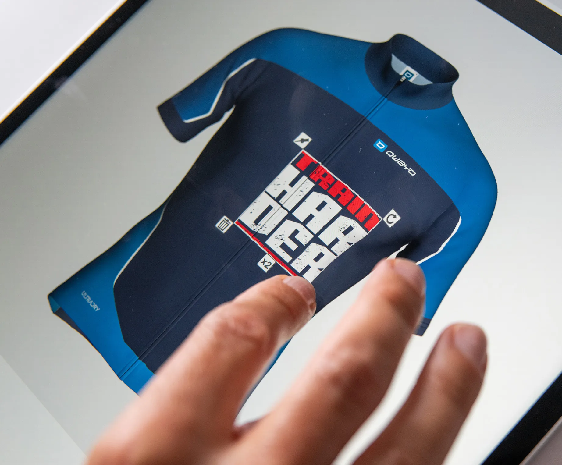 Una mano toca una pantalla que muestra una camiseta deportiva personalizada de color azul oscuro y azul con el texto 'TRAIN HARDER' y el logotipo de owayo.