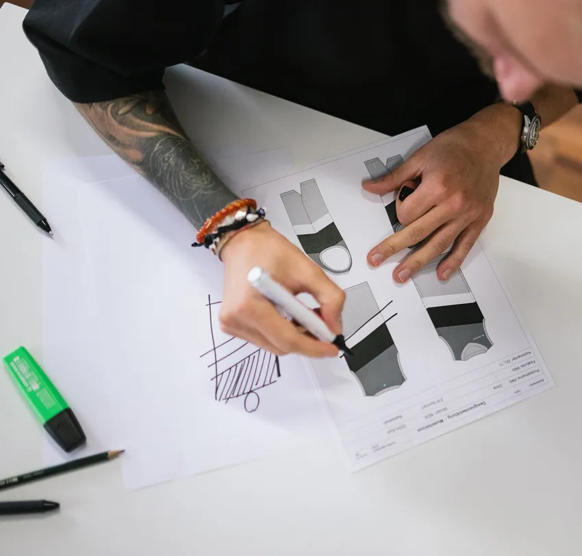 Una persona con un brazo tatuado dibuja diseños para ropa deportiva personalizada con un marcador sobre una hoja de papel.