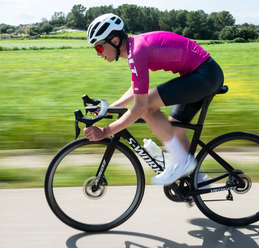 Un ciclista con un maillot rosa y pantalones cortos negros pedalea por una carretera a través de un paisaje verde, con un efecto de desenfoque de movimiento que resalta la velocidad.