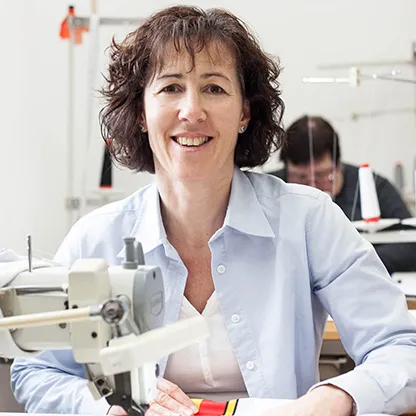 Una mujer sonríe a la cámara mientras trabaja en una máquina de coser en un taller, con otra persona visible en el fondo también trabajando en una máquina de coser.