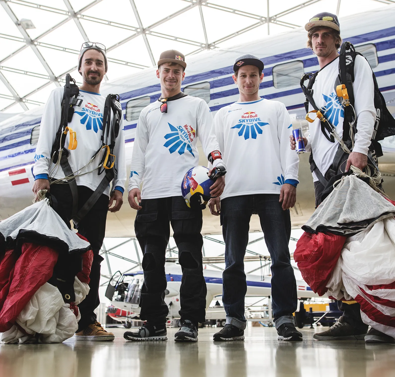 Cuatro hombres con camisetas blancas de manga larga con el logo del equipo Red Bull Skydive están de pie en un hangar frente a un avión.