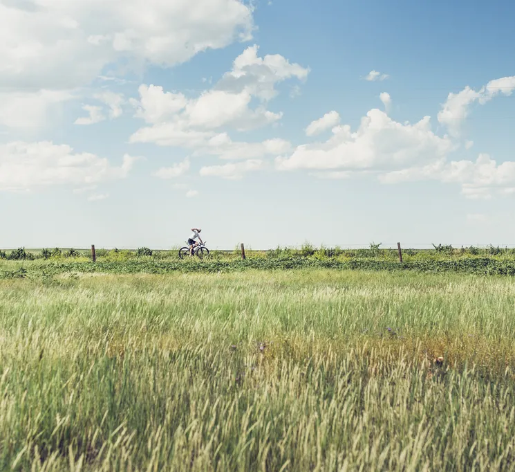 Un ciclista en un paisaje rural con hierba alta y cielo azul, simbolizando responsabilidad ecológica y actividades al aire libre.