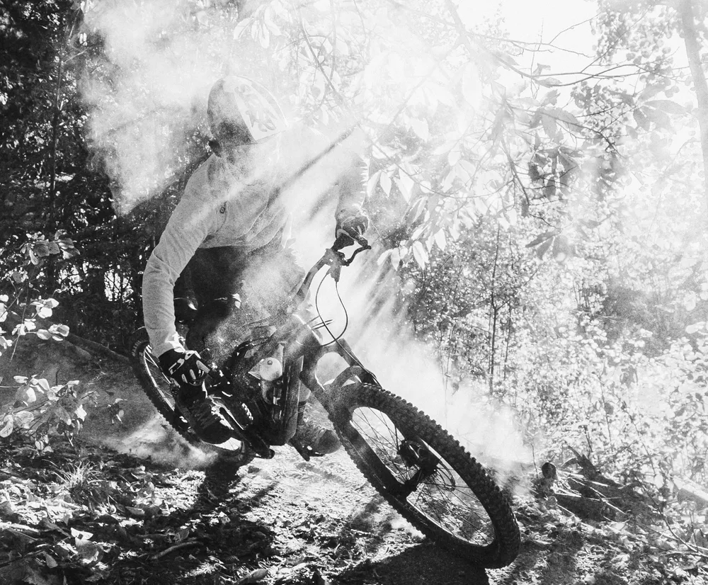 Un ciclista de montaña en movimiento en un sendero forestal, rodeado de polvo y rayos de luz, lleva una prenda de manga larga de owayo.