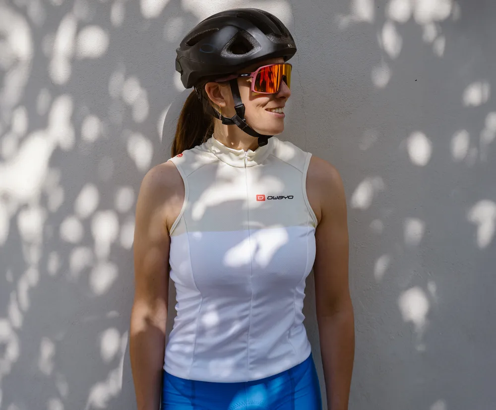 Una mujer con un jersey de ciclismo blanco sin mangas con el logo de owayo, un casco de bicicleta y gafas de sol, apoyada contra una pared con sombras de hojas.