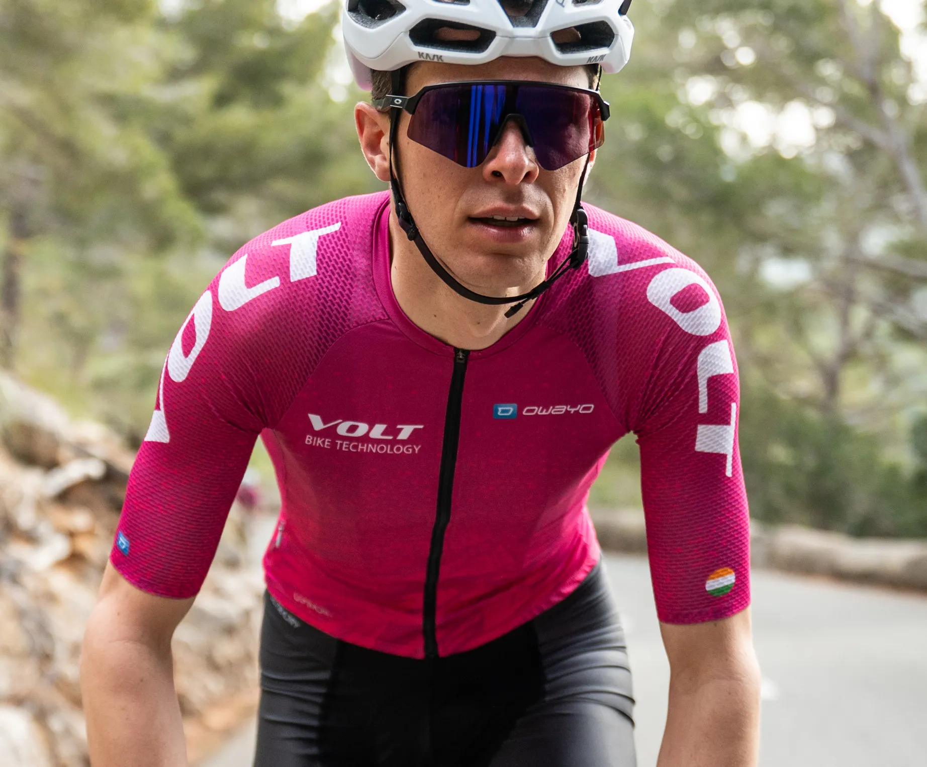 Un ciclista con una camiseta de ciclismo rosa de manga corta con el logo 'VOLT BIKE TECHNOLOGY' y el logo 'owayo' en el pecho, que pedalea por una carretera en una zona boscosa.
