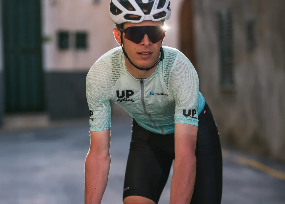 Un ciclista con una camiseta de ciclismo azul claro transpirable con los logotipos 'UP cycling' y 'owayo' en el pecho, en una postura deportiva en una calle.