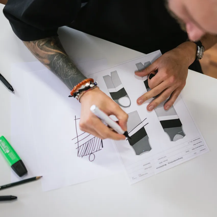 Una persona con un brazo tatuado está esbozando diseños para ropa deportiva en papel, rodeada de utensilios de escritura.