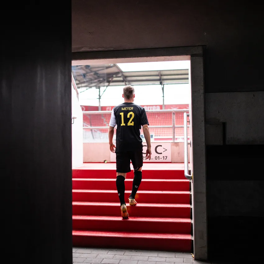 Un futbolista con una camiseta negra con el número 12 sube las escaleras rojas de un estadio.