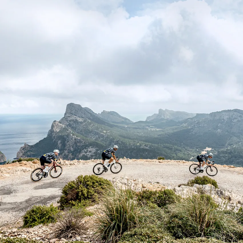 Tres ciclistas con ropa deportiva recorren un camino estrecho con vista a un paisaje costero.