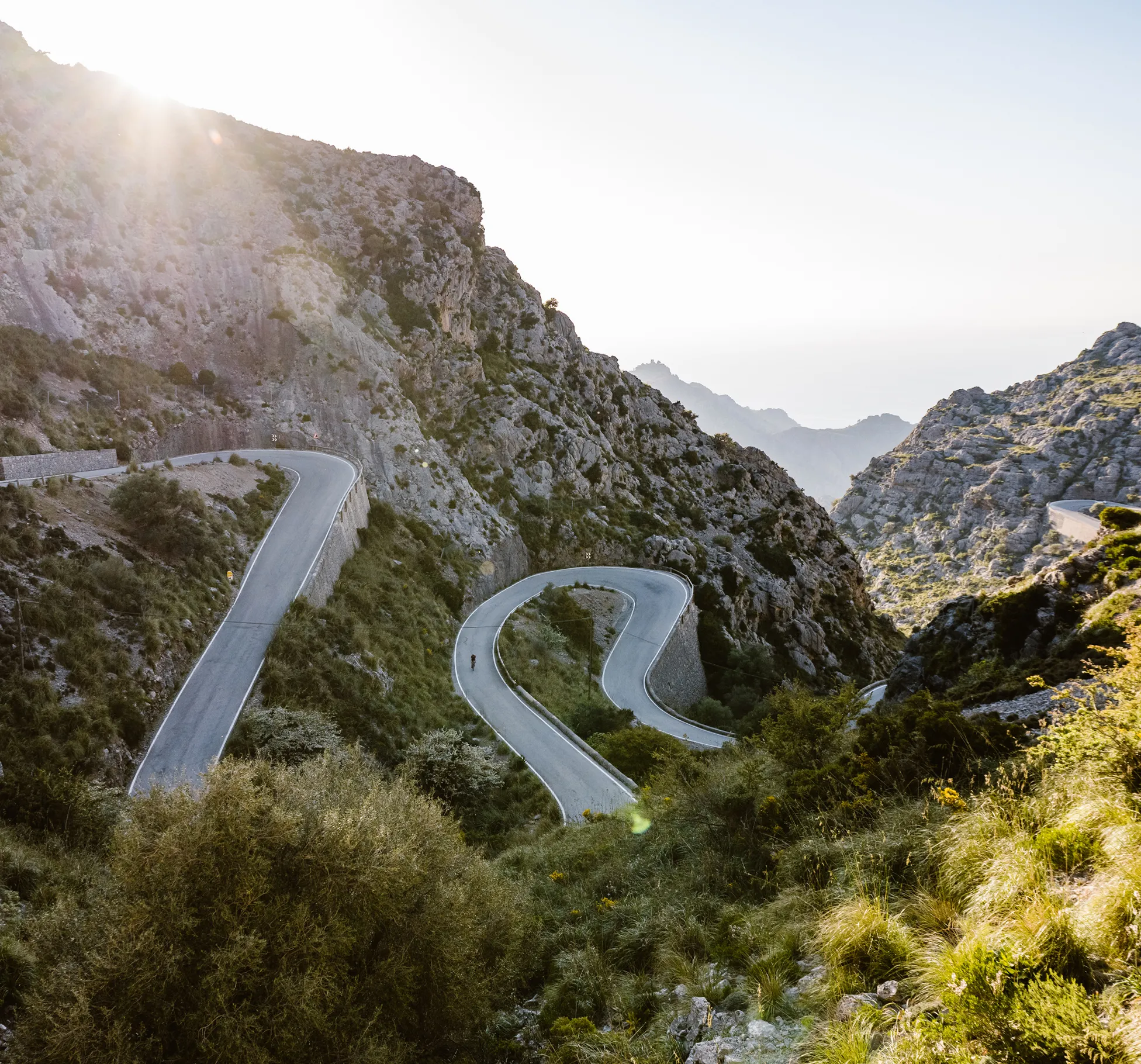 Una persona monta en bicicleta por una carretera de montaña sinuosa que serpentea a través de un paisaje rocoso, con la luz del sol brillando desde arriba.