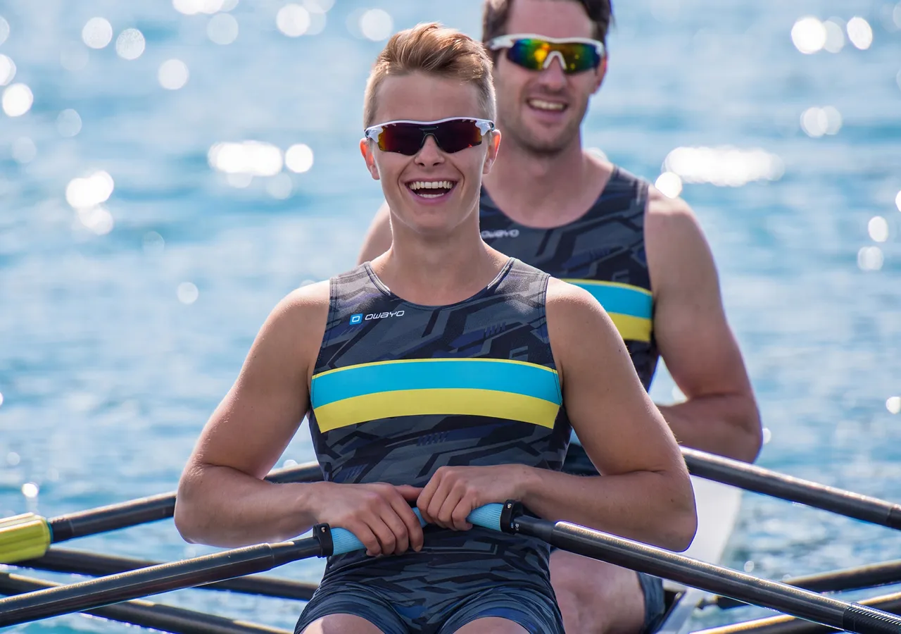 Dos atletas con ropa deportiva elegante están sentados en un bote de remo, sonriendo a la cámara mientras reman sobre agua brillante.