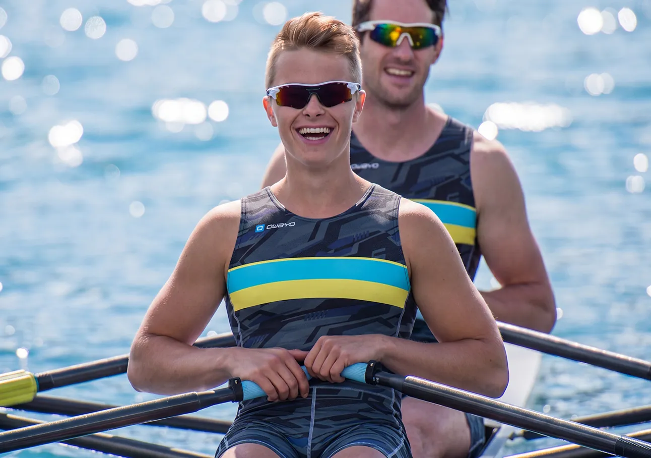 Dos remeros alegres con trajes deportivos de rayas azules y amarillas sentados en un bote sobre agua brillante.