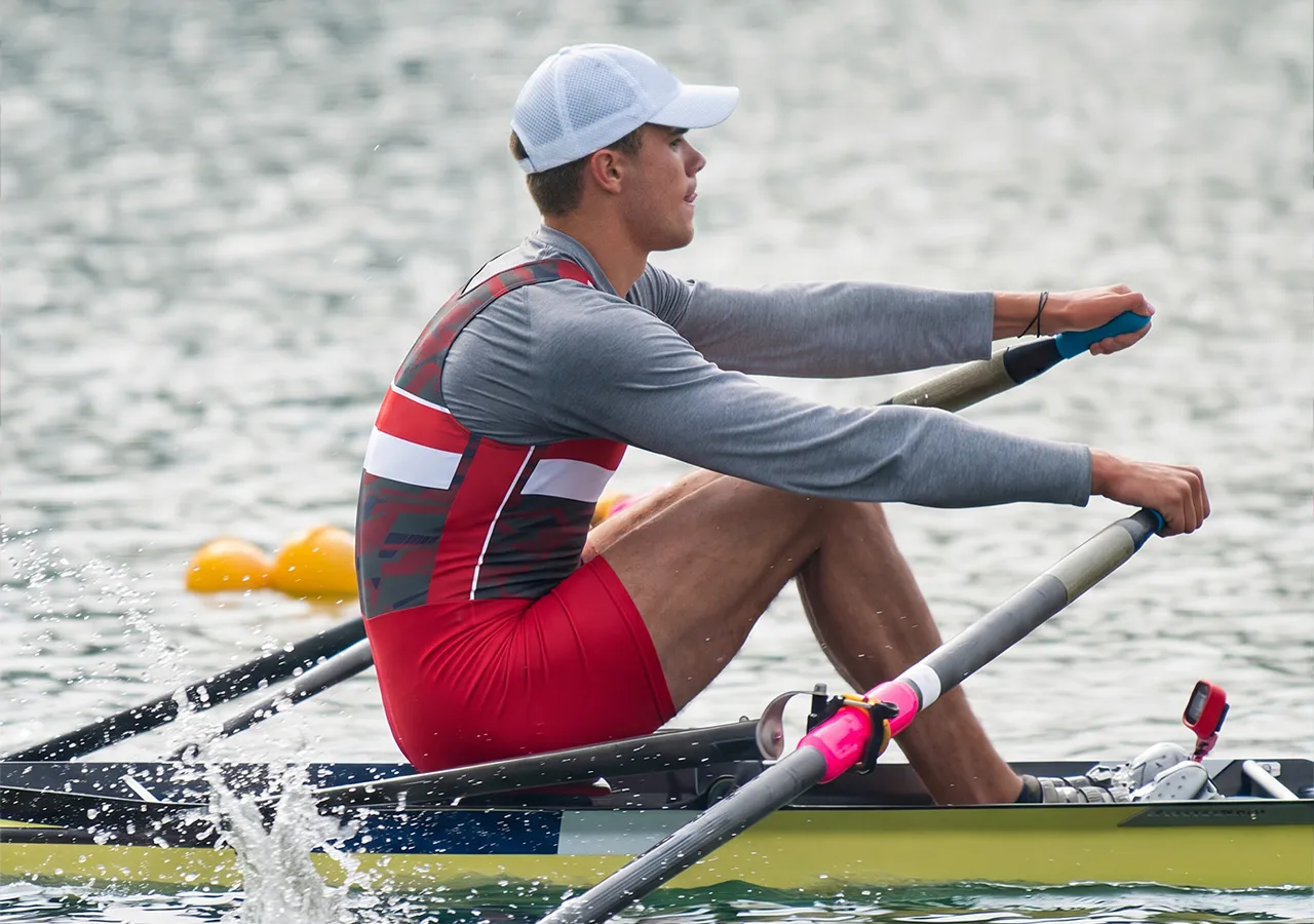 Un remero con un atuendo deportivo rojo y gris está remando en un cuerpo de agua.