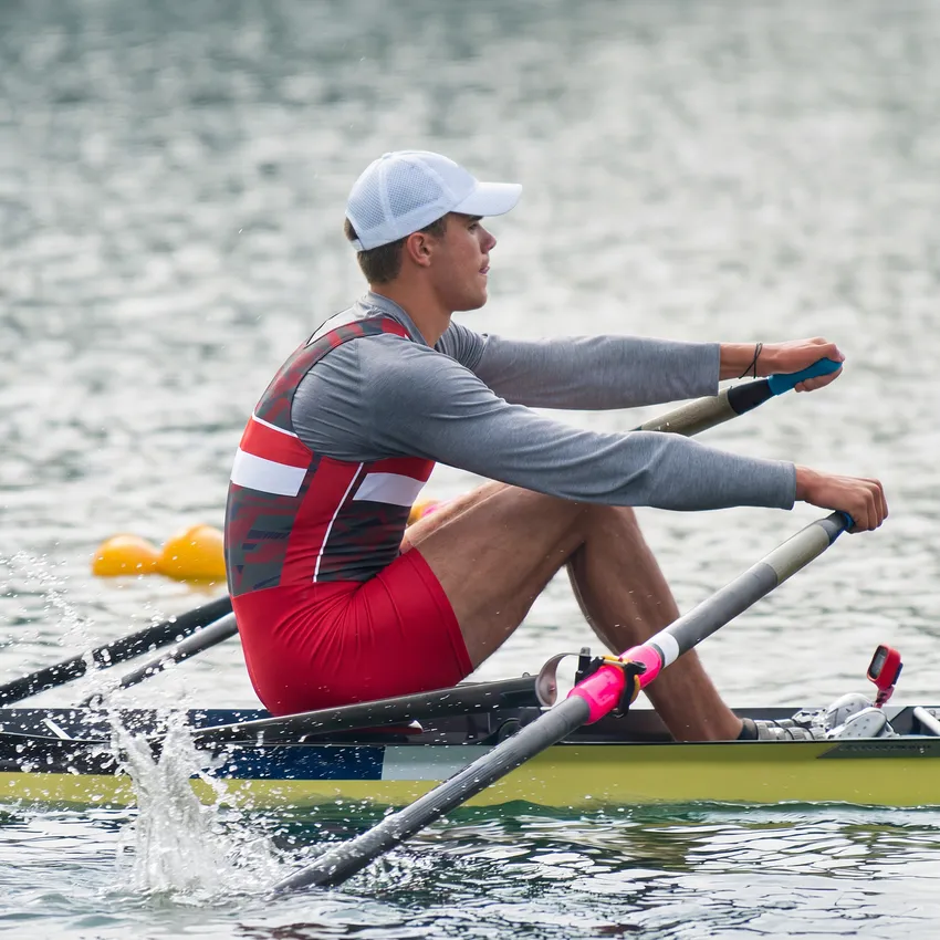 Un remero con un traje deportivo rojo y gris está remando en un cuerpo de agua.