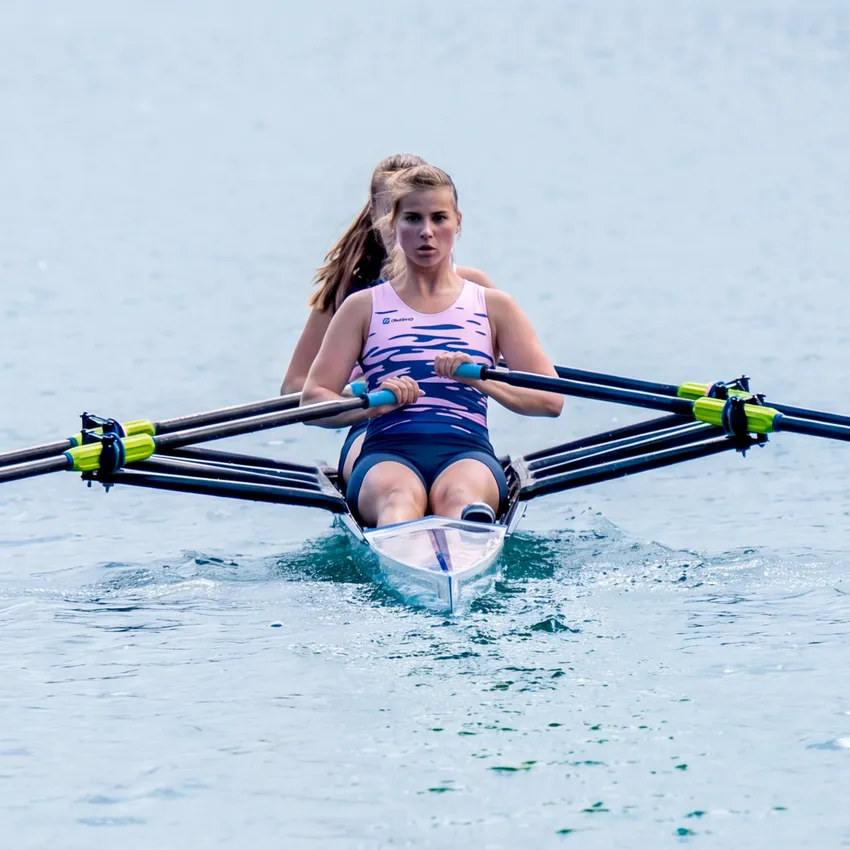 Dos remeros en un bote remando en aguas tranquilas, con la remera de adelante vistiendo un atuendo deportivo rosa.