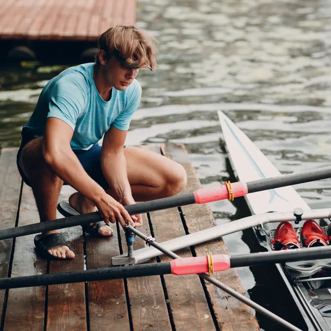 Un joven en una camiseta deportiva azul está sentado en un muelle ensamblando remos para un bote, con agua y un bote visibles al fondo.