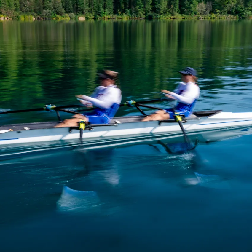 Dos remeros con ropa deportiva azul haciendo movimientos potentes en un lago tranquilo.