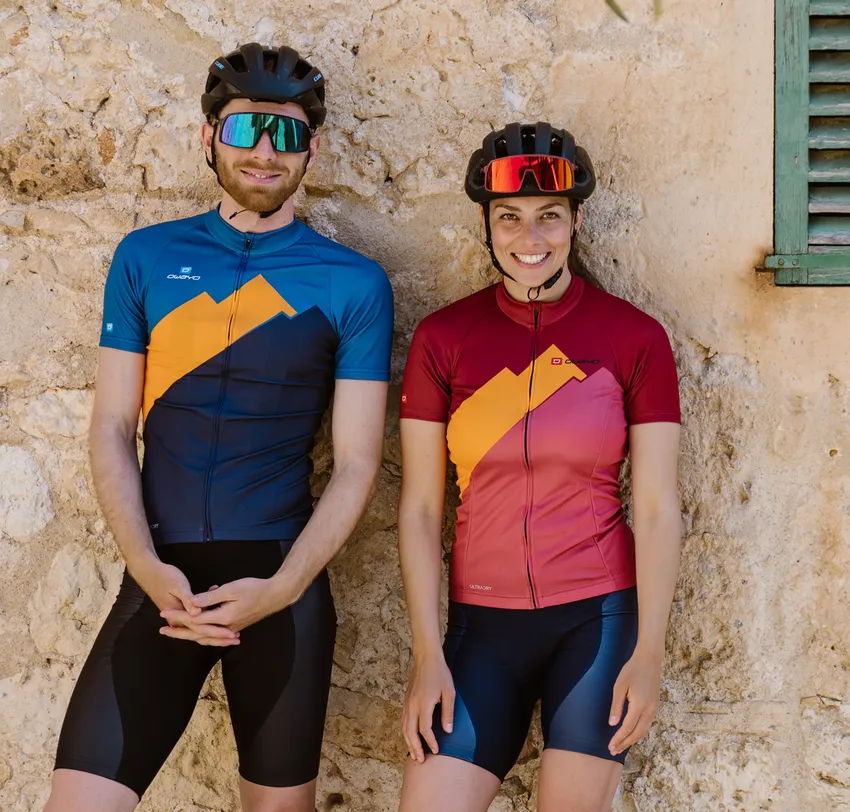 Un hombre y una mujer con camisetas de ciclismo personalizadas de owayo, de pie frente a una pared de piedra.