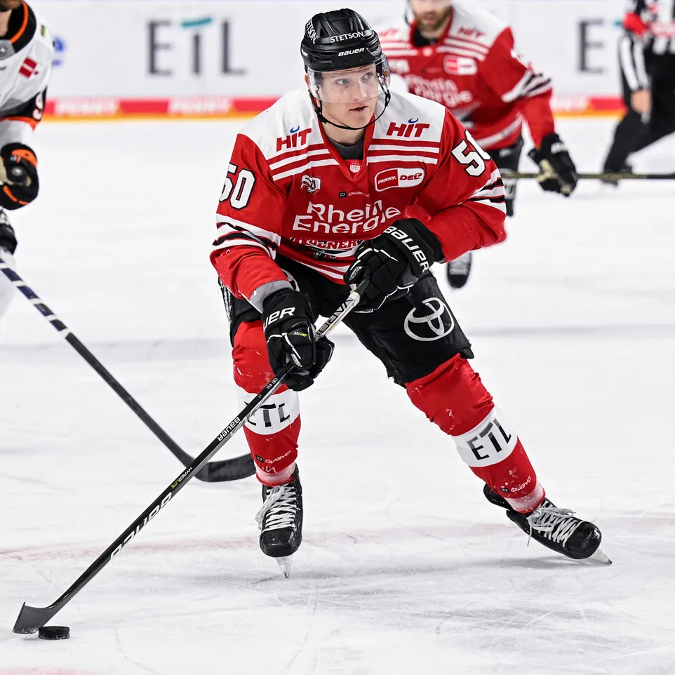 Un jugador de hockey sobre hielo con un jersey rojo con el número 50 y la inscripción 'Rhein Energie', en la pista de hielo, controlando un disco con su palo.
