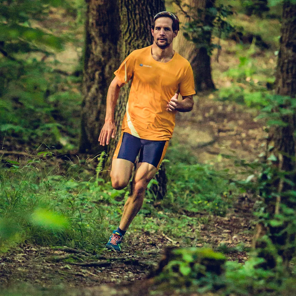 Un hombre corre por un sendero forestal con una camiseta de running naranja y shorts de running azul oscuro.