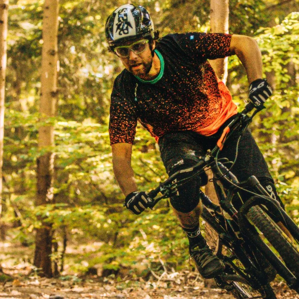 Un ciclista de montaña con una camiseta negra con puntos rojos y pantalones cortos negros monta en bicicleta por un bosque.