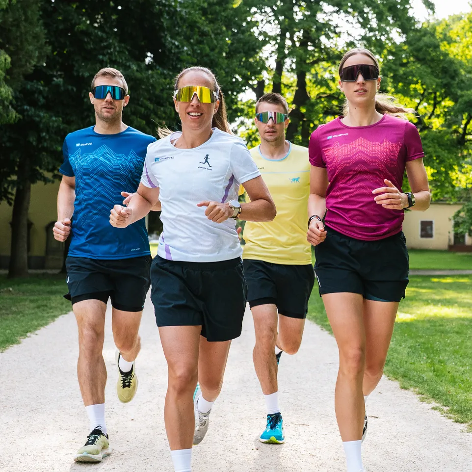 Cuatro corredores con camisetas deportivas coloridas y pantalones cortos corren por un camino en un parque.