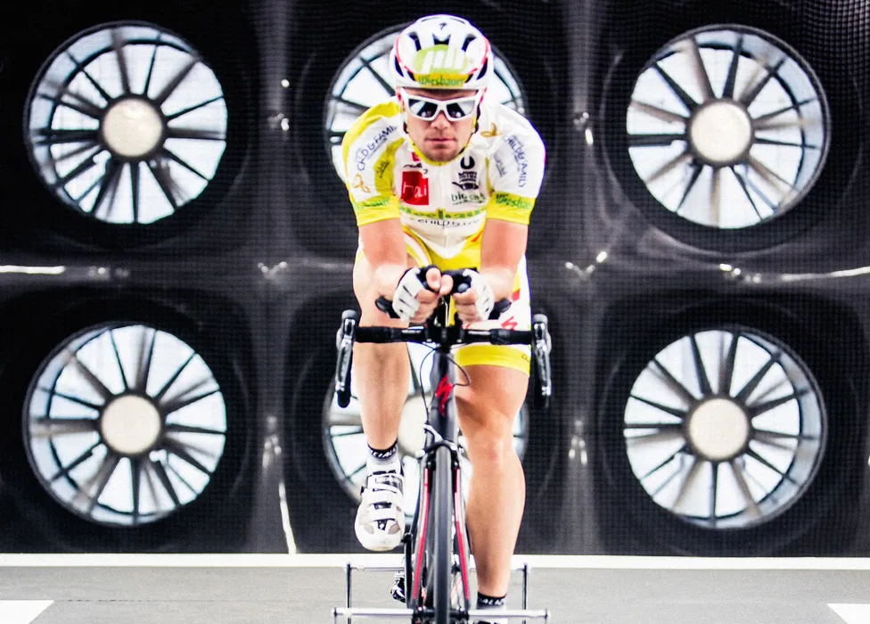 Un ciclista con maillot amarillo y blanco y casco pedalea en una bicicleta de entrenamiento frente a un fondo con grandes ventiladores.