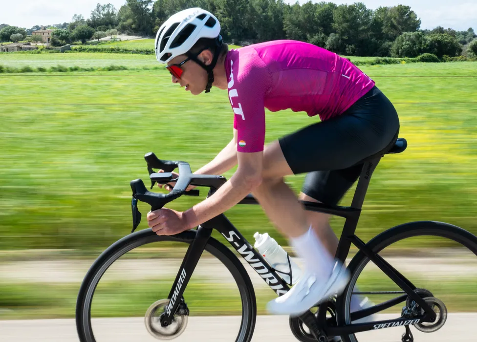 Un ciclista con maillot rosa y shorts negros monta una bicicleta de carretera en una carretera rural, con un fondo verde borroso que sugiere velocidad.
