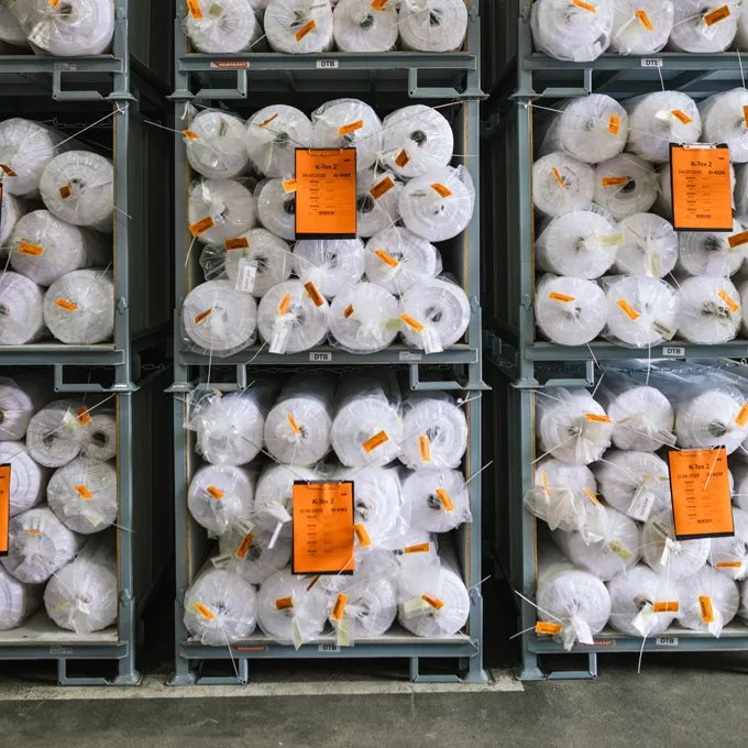 Un almacén con estanterías llenas de grandes rollos de tela blanca envueltos en plástico y con etiquetas naranjas.