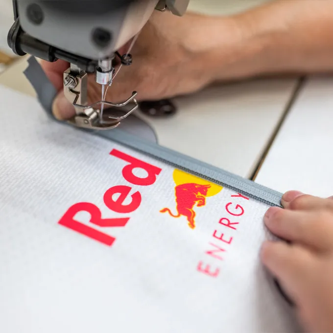 Un primer plano de una máquina de coser que cose una prenda deportiva blanca con el logotipo de Red Bull, mientras una persona sostiene el material.